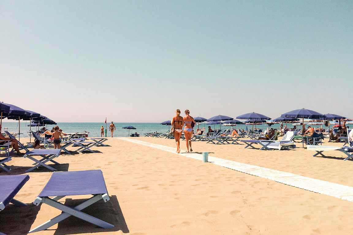 En sandstrand med blå liggestole og parasoller. Folk soler sig og går på stranden med en klar blå himmel i baggrunden. (AI-genereret tekst)
