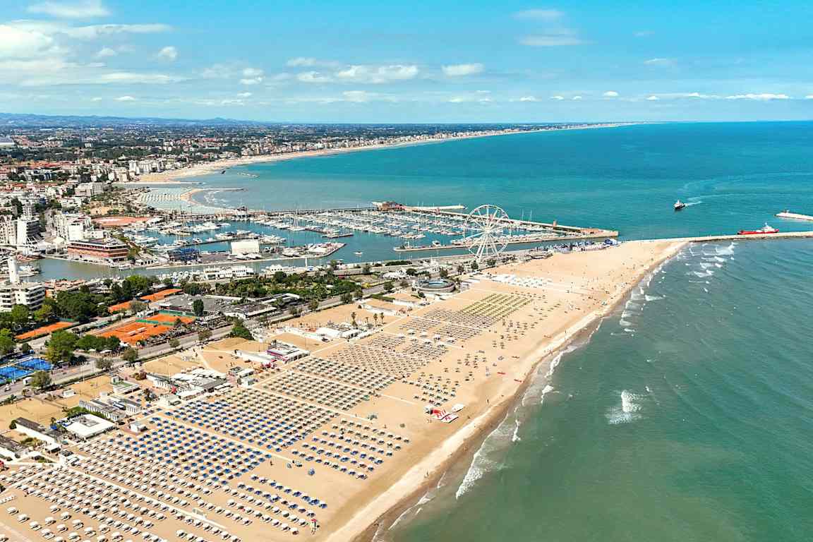 En udsigt over en bred sandstrand med række efter række af liggestole og parasoller. I baggrunden ses en marina og et forlystelsesområde-lignende miljø med et stort pariserhjul. Havet strækker sig til højre. (AI-genereret tekst)