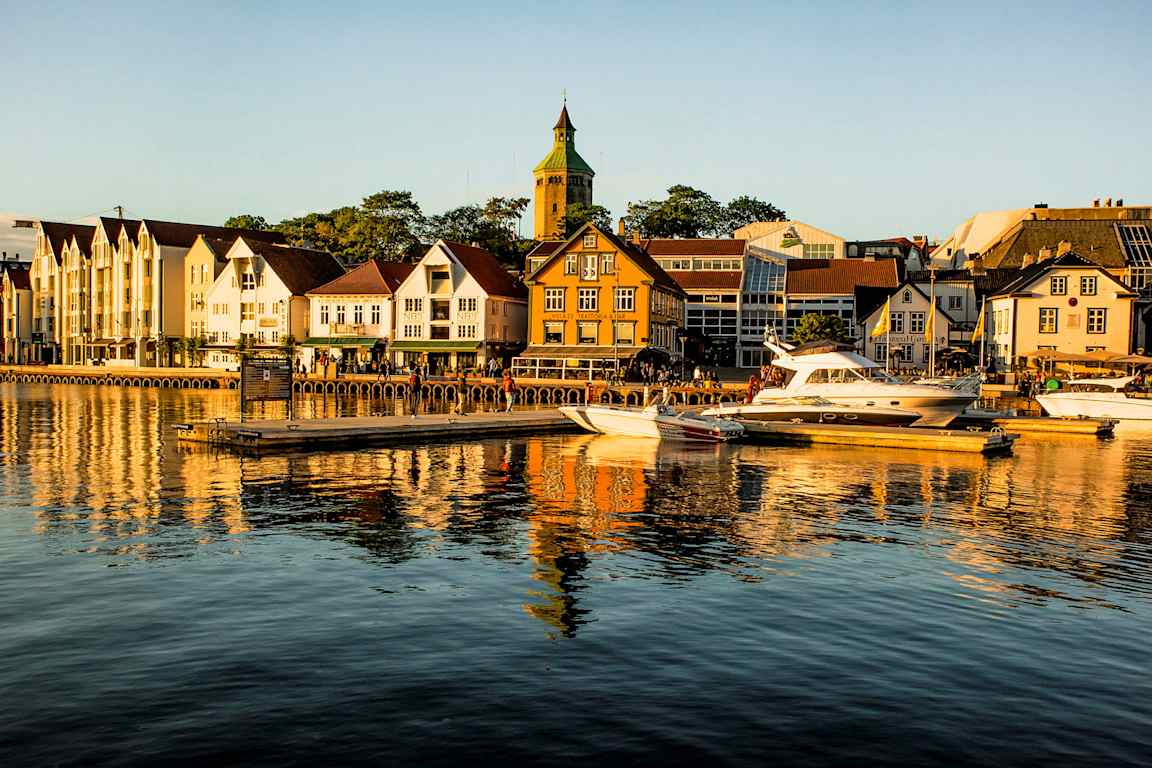 En havn med både liggende ved broen foran bygninger og en kirke i baggrunden. (AI-genereret tekst)