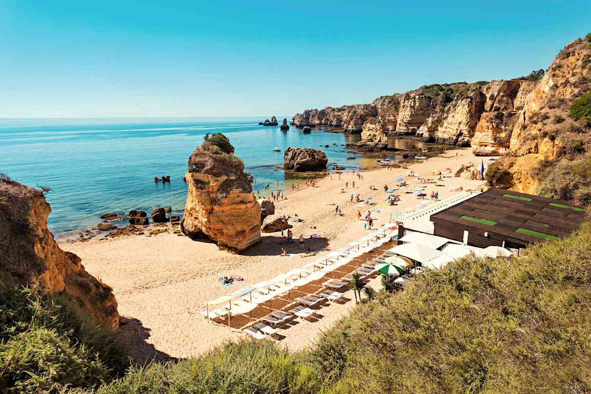 En sandstrand med liggestole og parasoller, omgivet af klippeformationer og et roligt hav. (AI-genereret tekst)