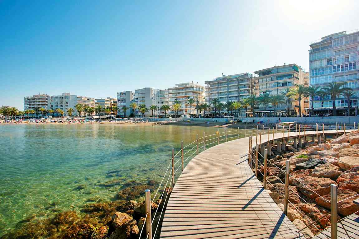 Billede af en strandpromenade med en gangbro, klart vand, palmer og bygninger i baggrunden. (AI-genereret tekst)