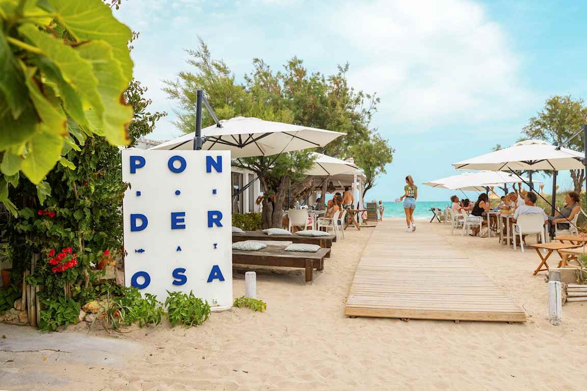 En restaurant ved stranden med borde og parasoller, en træsti fører mod vandet. (AI-genereret tekst)