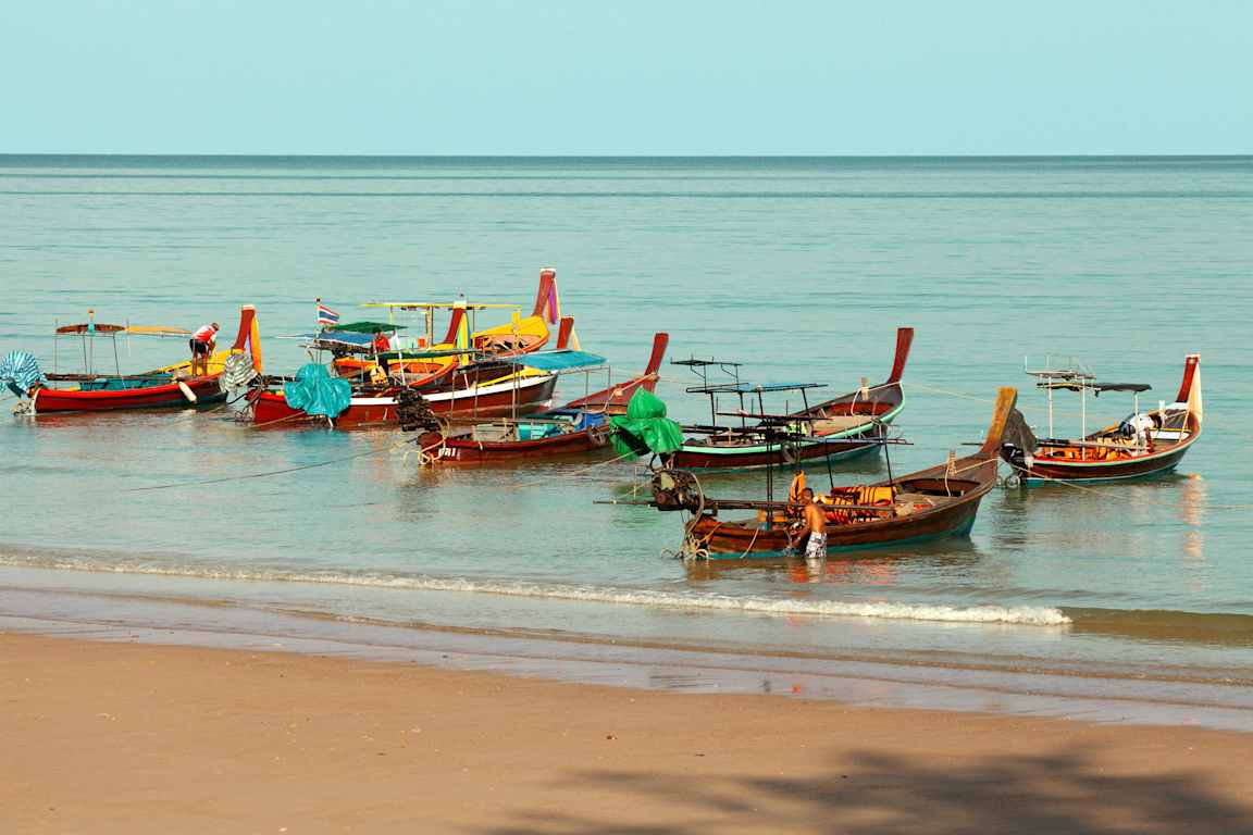 Flere farverige både ligger fortøjet i vandet nær en sandstrand. (AI-genereret tekst)
