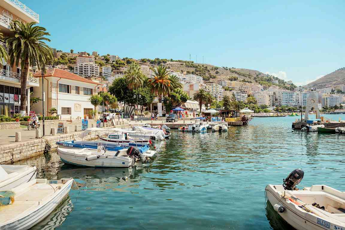 Både ligger fortøjet ved en kaj med bygninger og bakker i baggrunden. (AI-genereret tekst)