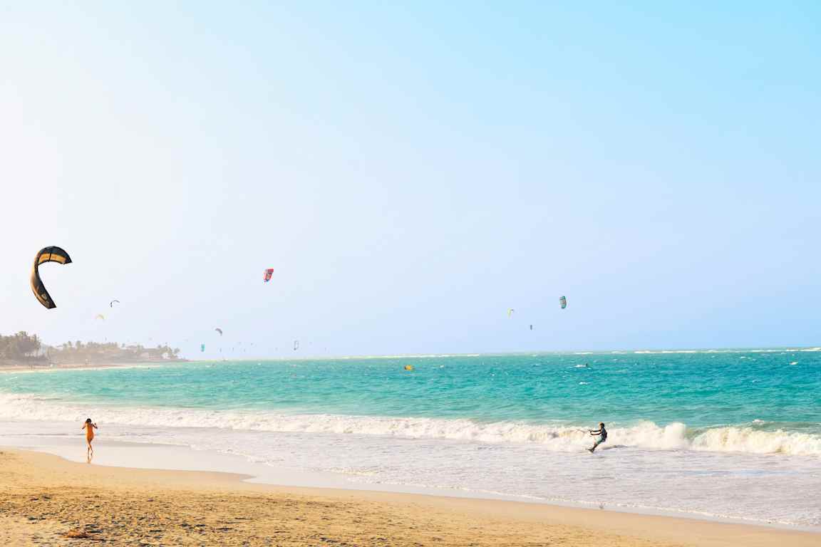 En strand med turkis hav og mange kitesurfere. (AI-genereret tekst)