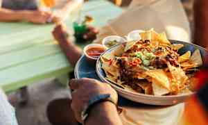 En person holder en tallerken med nachos, hvor forskellige dipsaucer er placeret rundt om tallerkenen. Scenen er udendørs ved et bord nær vand. (AI-genereret tekst)