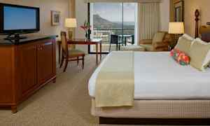 Et hotelværelse med en stor seng, et TV, et skrivebord og en stol. I baggrunden ses en balkon med udsigt over bjerge og hav. (AI-genereret tekst)