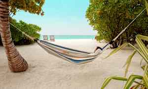 Person der hviler i en hængekøje på en sandstrand med træer og hav i baggrunden. (AI-genereret tekst)