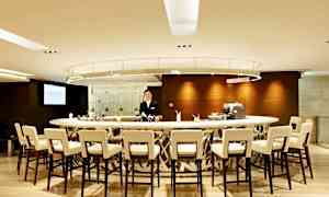 En moderne bar med en rund bardisk, omgivet af stole. En person står bag bardisken. (AI-genereret tekst)