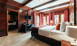 Hotelværelse med træindretning, en bred seng med hvide og farverige puder, røde gardiner og vinduer med udsigt. (AI-genereret tekst)