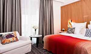 Hotelværelse med seng, mønstret pude, rødt tæppe, stol og bord med blomster. (AI-genereret tekst)