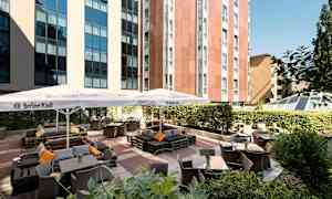 Billede af et udendørs serveringsområde med borde, stole og parasoller ved en bygning. (AI-genereret tekst)