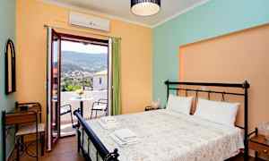 Et soveværelse med en dobbeltseng, to foldede håndklæder og et skrivebord ved siden af. Bag en åben dør ses en terrasse med bord og stole samt udsigt over et bjerglandskab. (AI-genereret tekst)