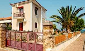 En to-etagers bygning med altan omgivet af en stenmur og port, ved siden af en palme og blomstrende buske. (AI-genereret tekst)