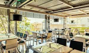 En restaurant med flere dækkede borde og stole, med vinduer mod en udsigt over byen. (AI-genereret tekst)