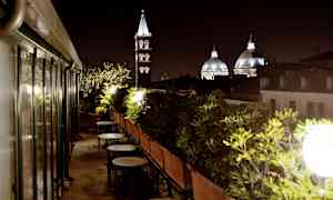 En tagterrasse med borde og stole omgivet af grønne planter, med kirketårne og kupler synlige i baggrunden om aftenen. (AI-genereret tekst)
