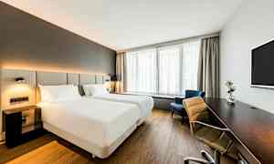 Et lyst hotelværelse med to enkeltsenge, et skrivebord, et tv og et vindue med gardiner. (AI-genereret tekst)