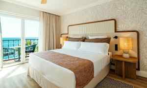 Et hotelværelse med en stor seng, to natborde med lamper og en balkon med udsigt over havet. (AI-genereret tekst)