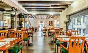 Et interiør af en restaurant med tomme borde og stole, træbjælker i loftet og store vinduer mod en gade. (AI-genereret tekst)