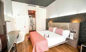 Et lyst hotelværelse med en dobbeltseng redt med hvidt sengetøj og lyserødt sengetæppe, et skrivebord med en stol og en dekorativ væg bag sengen. (AI-genereret tekst)