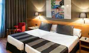 Et hotelværelse med to enkeltsenge, begge redt op med sort-hvide sengetæpper. På væggen hænger et farverigt maleri, og værelset har også et lille bord, en rød lænestol og to væglamper. (AI-genereret tekst)