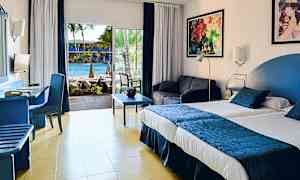 Et hotelværelse med en stor seng, siddeområde og udsigt over en pool gennem glasdøre. (AI-genereret tekst)