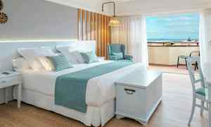 Et lyst hotelværelse med en stor dobbeltseng, stol og bord, samt en balkon med udsigt over havet. (AI-genereret tekst)