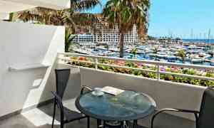 Udsigt fra en altan med et glasbord og to stole, der vender ud mod en marina og havet. (AI-genereret tekst)