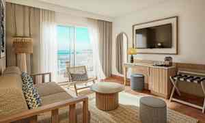 Et lyst og moderne hotelværelse med udsigt over havet. Der er en sofa, et sofabord, en stol og et fjernsyn på væggen. (AI-genereret tekst)