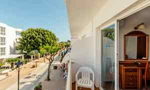 Udsigt fra en altan med en hvid plaststol. Gaden nedenfor har træer og bygninger. (AI-genereret tekst)