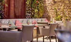 En udendørs serveringsområde med siddepladser og dekorerede vægge, omgivet af grønne områder. (AI-genereret tekst)