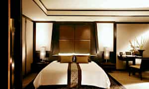 Et moderne soveværelse med en stor seng i midten, omgivet af elegante mørke træpaneler og varme lyskilder. (AI-genereret tekst)