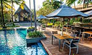 En udendørs pool omgivet af en træterrasse med borde og stole. Palmer og tropiske planter omgiver området. (AI-genereret tekst)