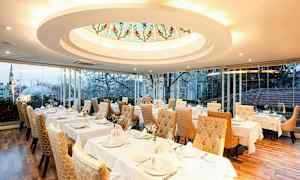 En elegant restaurant med dækkede borde og gyldne polstrede stole. Et stort vindue viser udsigten over en by med træer og bygninger. (AI-genereret tekst)