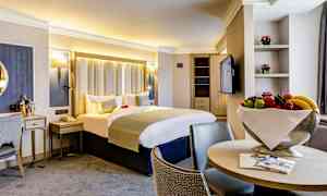 Lyst hotelværelse med dobbeltseng, stole og frugtskål. (AI-genereret tekst)