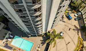 En høj bygning set oppefra med altaner, der vender udad. Nedenfor er der en pool, liggestole og parkerede biler. (AI-genereret tekst)