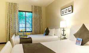 Et hotelværelse med to brede senge, hver med en pude og et tæppe. Et vindue med gardiner og udsigt til træer. (AI-genereret tekst)