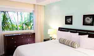 Et soveværelse med en seng, to malerier, et vindue med udsigt til frodige palmer og en kommode. (AI-genereret tekst)