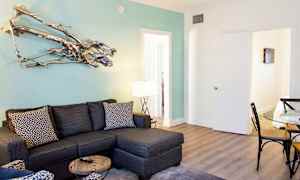 En moderne stue med en grå hjørnesofa, et glasbord og et siddeområde med mønstrede puder. En dekorativ metalskulptur hænger på en lyseblå væg. (AI-genereret tekst)