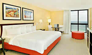 Et hotelværelse med en stor seng med hvidt sengetøj og en orange sengbund. Værelset har to malerier over sengen, et rundt bord med to stole, en orange siddepuf og et vindue med havudsigt. (AI-genereret tekst)