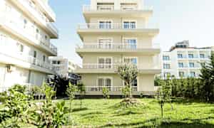 En flerfamilieejendom i lys farve med en have med græsplæne og små træer i forgrunden. (AI-genereret tekst)