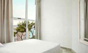 Et soveværelse med en hvid seng nær et stort vindue med udsigt over havet og nærliggende bygninger. (AI-genereret tekst)