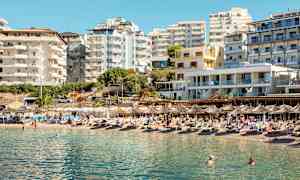 En strand med liggestole og parasoller foran flere moderne bygninger. (AI-genereret tekst)