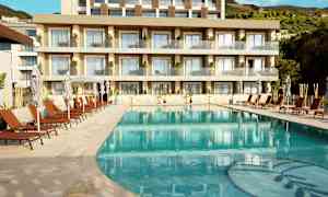 Billede af en swimmingpool med liggestole og parasoller foran et moderne hotel med balkoner. (AI-genereret tekst)