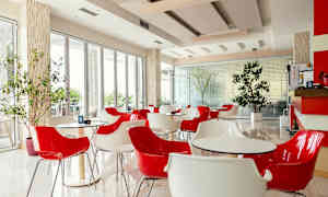 En moderne café med røde og hvide stole samt borde, beliggende i et lyst rum med store vinduer og indendørs planter. (AI-genereret tekst)