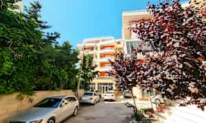 Billedet viser en boligbygning med parkerede biler foran og buske samt træer omkring. (AI-genereret tekst)