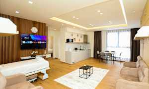 Et moderne hotelværelse med åbent gulvplan, der inkluderer en sovesofa, TV, køkkenområde, spisebord og store vinduer med gardiner. (AI-genereret tekst)