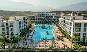 Et moderne hotelanlæg med en stor pool og omkringliggende liggestole, beliggende i en frodig dal med bjerge i baggrunden. (AI-genereret tekst)