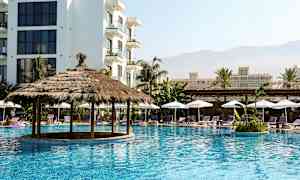 En pool med en stråtagspavillon omgivet af liggestole og parasoller, med flere bygninger i baggrunden. (AI-genereret tekst)