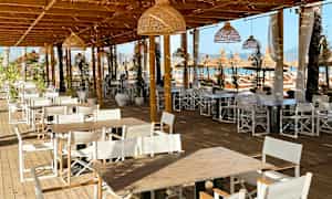 Billede af en udendørs spiseplads med træborde og hvide stole under en træpergola. (AI-genereret tekst)